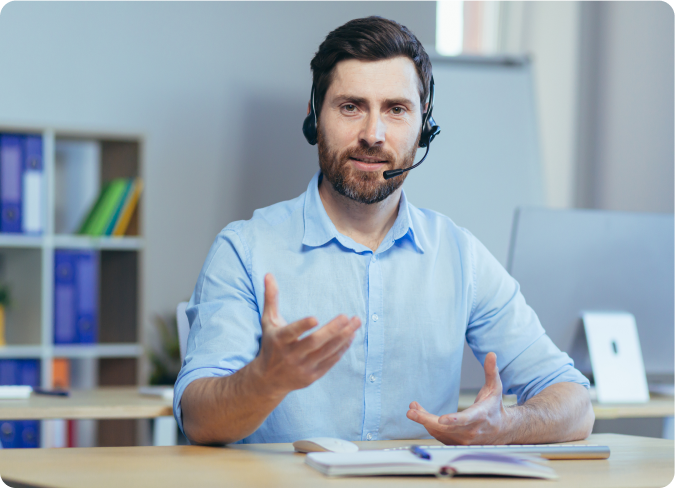 A tutor wearing a headset, sitting at a desk with books and a laptop, gesturing while speaking